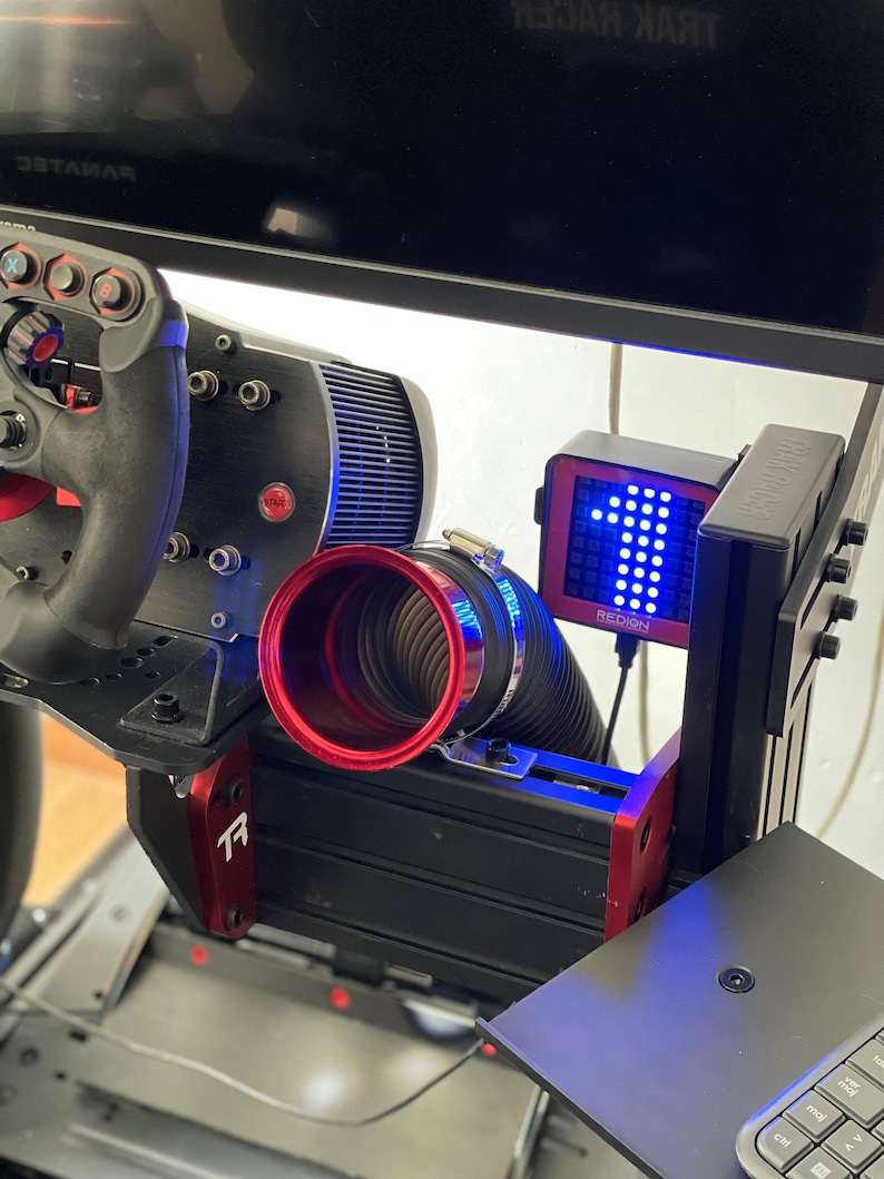 May include: A black and red racing simulator setup with a steering wheel, pedals, and a monitor. The setup includes a red and black frame, a digital display with the word "REDIGN", and a black keyboard.