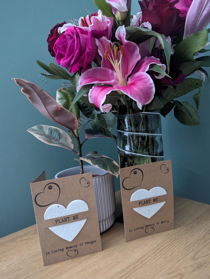 May include: Two brown cards with white heart-shaped seed paper that says "Plant Me" on them. The cards are on a wooden surface with a bouquet of pink and purple flowers in a glass vase behind them. The text on the cards reads "In Loving Memory of Fergus" and "In Loving Memory of Molly".