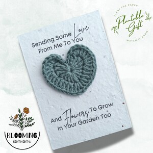 May include: A white card with the text "Sending Some Love From Me To You" and "And Flowers To Grow In Your Garden Too". A crocheted teal heart is centered on the card. The card is a plantable gift.