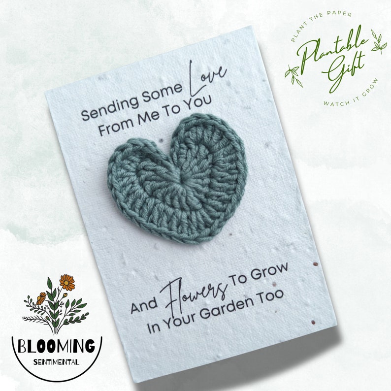 May include: A white card with the text "Sending Some Love From Me To You" and "And Flowers To Grow In Your Garden Too". A crocheted teal heart is centered on the card. The card is a plantable gift.