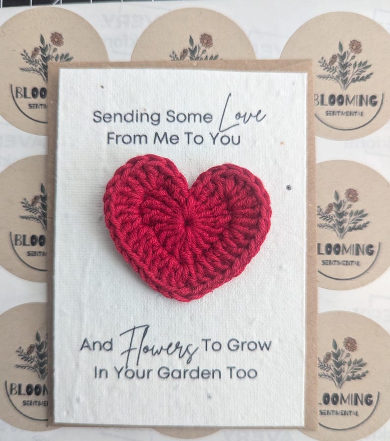 May include: A handmade greeting card with a red crocheted heart. The card reads "Sending Some Love From Me To You" and "And Flowers To Grow In Your Garden Too". The background includes circular stickers with floral designs.