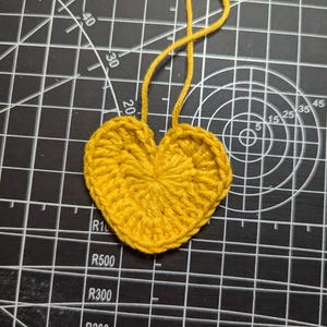 May include: A handmade, yellow crocheted heart with a loop of yarn attached at the top. The heart is made of tightly woven yarn and is set against a black cutting mat with white grid lines and measurements.