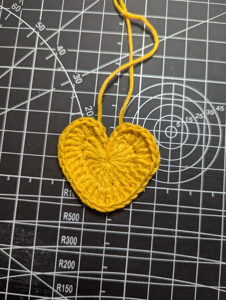 May include: A handmade, yellow crocheted heart with a loop of yarn attached at the top. The heart is made of tightly woven yarn and is set against a black cutting mat with white grid lines and measurements.