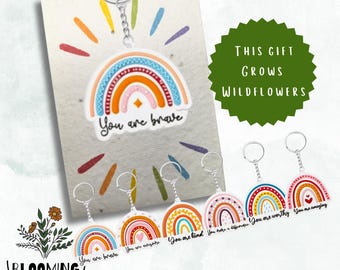 May include: A set of 7 rainbow-colored keychains with different positive messages, such as "You are brave", "You are worthy", and "You are amazing". The keychains are displayed on a white background with a card that reads "This gift grows wildflowers".