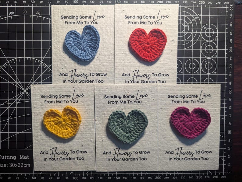 May include: A collection of handmade greeting cards, each featuring a crocheted heart in various colors: blue, red, yellow, green, and purple. The cards have the text "Sending Some Love From Me To You" and "And Flowers To Grow In Your Garden Too."