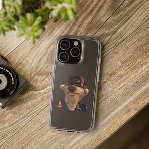 May include: Clear phone case featuring a cartoon character wearing a Red Bull Racing hat. The case has a clear border and a dark gray background. The phone case is on a wooden surface.