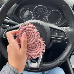 May include: Two pink crocheted coasters with fringe. The coasters are held in a hand in a car.