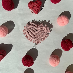 May include: A pink heart-shaped macrame keychain with a white plastic clasp. The keychain is surrounded by pink and red pom-poms.