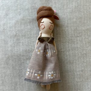 May include: A handmade doll with a brown bun hairstyle and a small pink flower. The doll wears a tan dress with white and yellow floral embroidery and a grey fringe hem. The doll's face has closed eyes and rosy cheeks.