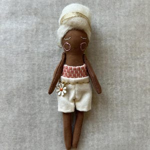 May include: A handmade doll with brown skin, wearing a white head wrap with a gold band, a pink and white patterned top, and white shorts. A gold chain with a daisy charm hangs from the shorts. The doll's arms and legs are brown.