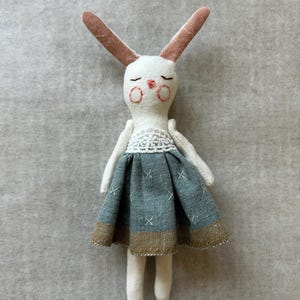 May include: A handmade white bunny doll with long, pink-lined ears. The doll wears a blue and brown skirt with a white lace top. The bunny has closed eyes and rosy cheeks, a charming and whimsical design.