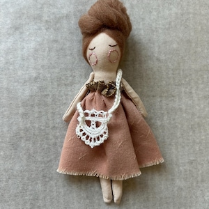 May include: A handmade cloth doll with a brown bun hairstyle, closed eyes, and a blush-coloured dress. The doll has a white crocheted purse and a gold-coloured collar. The doll's arms and legs are a light beige colour.