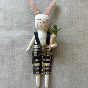 May include: A handcrafted white bunny doll with pink inner ears and rosy cheeks. The bunny wears plaid overalls with black braces and holds a small carrot. The doll is made of fabric and has a whimsical, handcrafted appearance.