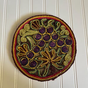 May include: Circular trivet with a grape design. The trivet features a brown border with a red, yellow, and green inner border. The grape design is made of purple, yellow, and green woven material. The trivet is likely used for hot dishes.