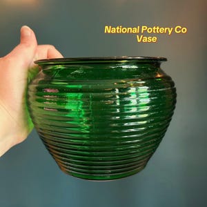 May include: A green glass vase with horizontal ribbed detailing. The vase has a wide, rounded shape and a dark rim. The text "National Pottery Co Vase" is displayed in yellow letters.