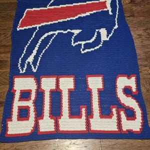 以下が含まれることがあります： 白と赤の Buffalo Bills のロゴと、赤色のアウトラインで囲まれた白い文字で「BILLS」と書かれた、青い編み物の毛布です。