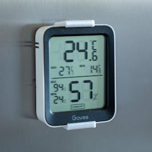 Könnte beinhalten: Ein weißes digitales Thermometer und Hygrometer mit einem schwarzen Display zeigt eine Temperatur von 24,6 Grad Celsius und eine Luftfeuchtigkeit von 57 %. Das Display zeigt auch die maximale Temperatur von 27 Grad Celsius und die minimale Temperatur von 14,5 Grad Celsius an. Das Gerät ist an einer silbernen Oberfläche montiert.