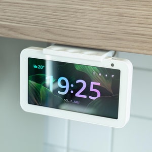 Könnte beinhalten: Ein weißes Smart Display mit einem schwarzen Bildschirm, der die Zeit 19:25 und die Temperatur 20 Grad anzeigt. Das Display ist unter einem Holzschrank montiert.
