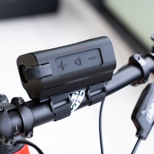 K&ouml;nnte beinhalten: Ein schwarzer Bluetooth-Lautsprecher, der mit schwarzen B&auml;ndern an einem Fahrradlenker befestigt ist. Der Lautsprecher hat Lautst&auml;rkeregler und eine Wiedergabe-/Pause-Taste.