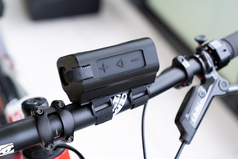 K&ouml;nnte beinhalten: Ein schwarzer Bluetooth-Lautsprecher, der mit schwarzen B&auml;ndern an einem Fahrradlenker befestigt ist. Der Lautsprecher hat Lautst&auml;rkeregler und eine Wiedergabe-/Pause-Taste.