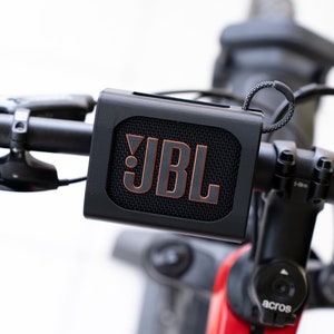 K&ouml;nnte beinhalten: Ein schwarzer JBL-Tragbarer Lautsprecher, der an den Lenker eines Fahrrads montiert ist.