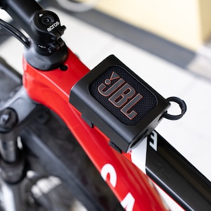 K&ouml;nnte beinhalten: Ein schwarzer JBL-Tragbarer Lautsprecher, der an einem roten und schwarzen Fahrradrahmen befestigt ist. Der Lautsprecher hat ein schwarzes Mesh-Gitter und ein orangefarbenes JBL-Logo.