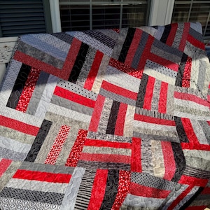 May include: A handmade quilt with a patchwork design in red, black, and gray. The quilt features a variety of patterned fabrics, including stripes, polka dots, and geometric shapes. The quilt is likely made of cotton and is suitable for a bed or sofa.