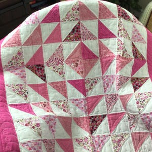 May include: A handmade quilt featuring a geometric pattern of pink and white triangles and squares. The quilt has a pink border with a decorative stitched pattern. The quilt is likely made of cotton fabric.