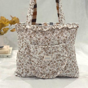 Può includere: Una borsa tote trapuntata in tessuto bianco con una piccola stampa floreale marrone. La borsa presenta un bordo arricciato lungo la parte superiore e sulla tasca frontale. Un'etichetta rettangolare sulla tasca frontale riporta FAYE'S Quilted Cottage.