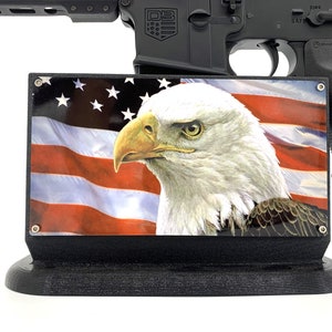 May include: A black rifle with a stand featuring an American flag with a bald eagle. The stand is made of black plastic and has a base.