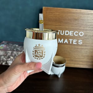Puede incluir: Calabaza de yerba mate blanca con borde dorado y el logo de la AFA en dorado. La calabaza tiene forma de cohete con una pajita bombilla a juego. También se ve una pequeña taza de mate a juego.