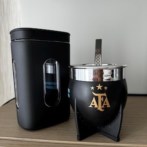 Peut inclure: Un récipient à maté noir avec un bord argenté et une paille en métal, à côté d'un récipient noir. Le récipient à maté présente un emblème doré avec trois étoiles et les lettres "AFA". Les articles sont sur une surface en bois.