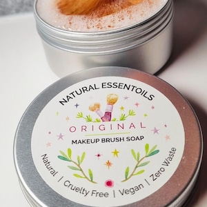 Op de afbeelding: Twee zilveren blikken met make-up kwast zeep. Eén blik is open met een make-up kwast erin, omringd door schuimende zeep. Het andere blik heeft een label met de tekst "NATURAL ESSENTOILS ORIGINAL MAKEUP BRUSH SOAP". Het label zegt ook "Natural | Cruelty Free | Vegan | Zero Waste".