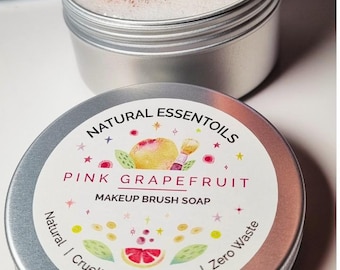 May include: Two silver tins of makeup brush soap. The top tin holds a makeup brush in a foamy solution. The bottom tin has a label that reads "NATURAL ESSENTOILS PINK GRAPEFRUIT MAKEUP BRUSH SOAP". The label also states "Natural | Cruelty Free | Vegan | Zero Waste".