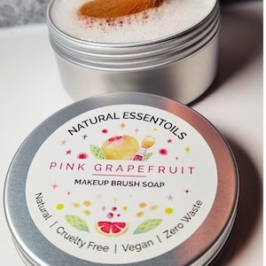 May include: A silver tin with a pink grapefruit design and the text "NATURAL ESSENTOILS PINK GRAPEFRUIT MAKEUP BRUSH SOAP Natural | Cruelty Free | Vegan | Zero Waste".