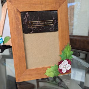 May include: A wooden picture frame with a brown finish. The frame has a decorative holly leaf and flower design in the bottom right corner. The frame has a small card inside with the text "Artisan Design & Frames" and the website address "www.artisan designandframes.com".