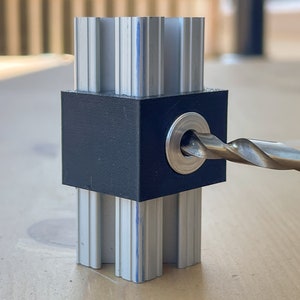 May include: A close-up of a drill bit going through a black plastic block with a metal ring. The block is attached to a silver aluminium extrusion.