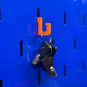 May include: An orange pegboard hook holds a set of black keys with silver key heads. The hook is mounted on a blue pegboard with vertical slots. The keys are attached to a silver key ring.