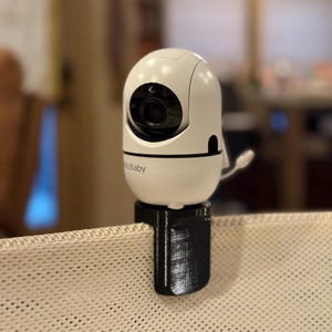 May include: A white baby monitor with a black base is attached to a crib. The monitor has a camera lens and the text "helloBaby" is visible. The monitor is mounted on a black clip.