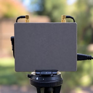 Può includere: Un dispositivo elettronico grigio rettangolare montato su una testa di treppiede nera. Il dispositivo presenta due connettori per antenna color oro sul bordo superiore, con cavi neri collegati ai lati e in alto, per un aspetto tecnico.