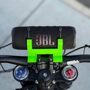 May include: A black JBL speaker mounted on a bicycle handlebar with a bright green holder. The speaker is secured with black zip ties. The JBL logo is red.