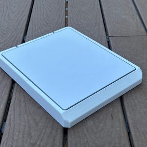 Könnte beinhalten: Eine weiße quadratische Zugangsklappe mit erhöhtem Rand aus Kunststoff. Die Klappe liegt auf einer braunen Holzoberfläche.