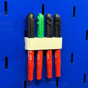 Può includere: Un portapenne bianco stampato in 3D con quattro pennarelli rossi, tre neri e uno verde. I pennarelli sono etichettati "FINE POINT 48-22-3100". Il supporto è montato su una pannello forato blu.