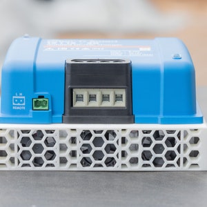 May include: A blue and white electronic device with a black fan and a white honeycomb-shaped grill. The device has a label that reads "Victron Energy" and "12/24V 100/50 3000VA 230V".