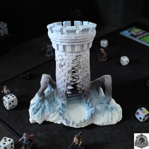 May include: A detailed, gray and white miniature castle tower with a cracked facade and icy base. The tower is surrounded by miniature figures and dice, suggesting a tabletop game setting. The tower is approximately 5 inches tall.