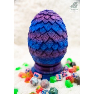 May include: A purple and blue dragon egg with a textured surface. The egg is sitting on a white surface with several colorful dice.
