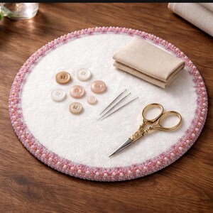 May include: A white circular sewing mat with a beaded pink and white border. The mat holds several buttons in shades of white and pink, two needles, a pair of gold scissors, and a folded piece of beige fabric. The items are arranged on a wooden surface.
