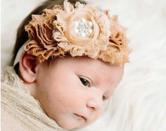 May include: A baby wearing a headband with a large, beige flower and a pearl-encrusted center. The flower is made of fabric and has a ruffled texture.