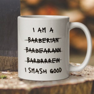 May include: White ceramic mug with black text that reads "I am a Barberian, Barbearann, Barbrrrein, I Smash Good". The mug is sitting on a tree stump next to an axe.