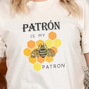 May include: A white t-shirt with a graphic of a bee in a honeycomb. The text "PATRÓN IS MY PATRÓN" is printed on the shirt.